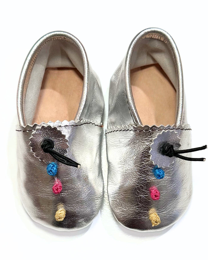A pair of metallic silver leather baby booties with scalloped edges, black drawstrings, and colorful embroidered accents in blue, pink, yellow, and gold on a white background.