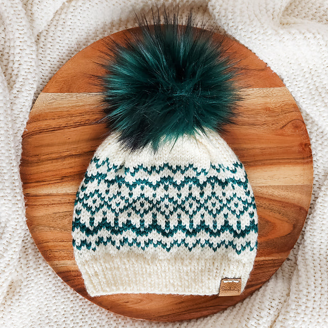 Finished Snow Days beanie in cream and teal Fair Isle snowflake pattern with dark teal faux fur pom pom displayed on a wooden board