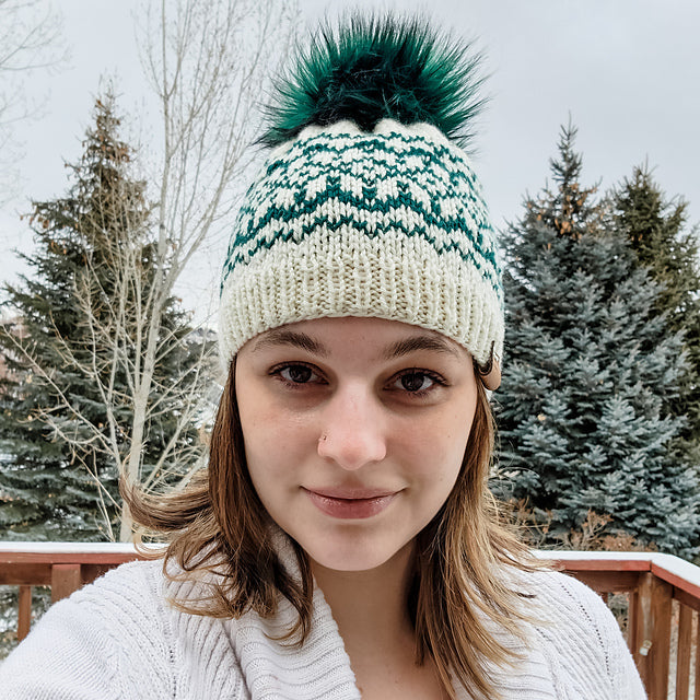 Woman wearing the Snow Days beanie in cream and teal Fair Isle pattern with teal faux fur pom pom outdoors in winter with snowy trees behind her