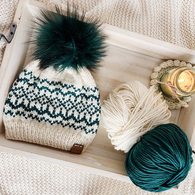 Snow Days beanie in cream and teal displayed in a wooden tray with cream and teal yarn balls and a lit candle on a crochet coaster