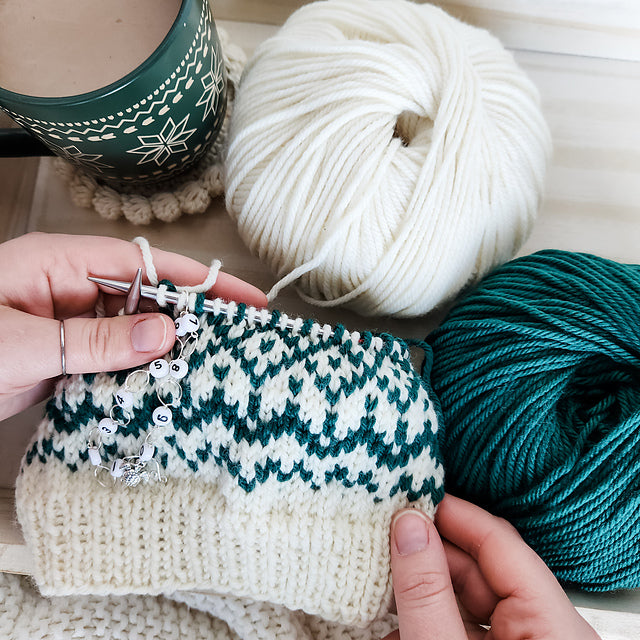 Hands knitting the Snow Days beanie in progress on circular needles with a row counter showing the teal and cream Fair Isle pattern with yarn balls and mug nearby
