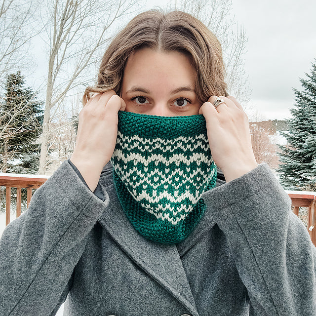 Woman wearing a teal and white stranded colorwork Snow Days Cowl pulled up over her nose outdoors in a snowy winter setting
