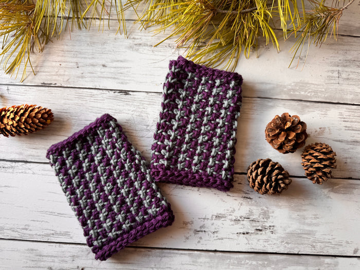Pair of Snowfall fingerless mitts in purple and gray Tunisian crochet colorwork laid flat on whitewashed wood with pine branches and pinecones