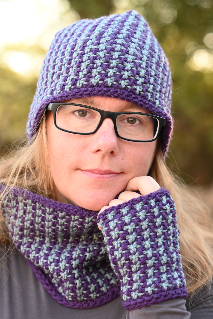 Woman wearing the complete Snowfall set including hat cowl and fingerless mitts in purple and gray with hand under chin showing the colorwork pattern