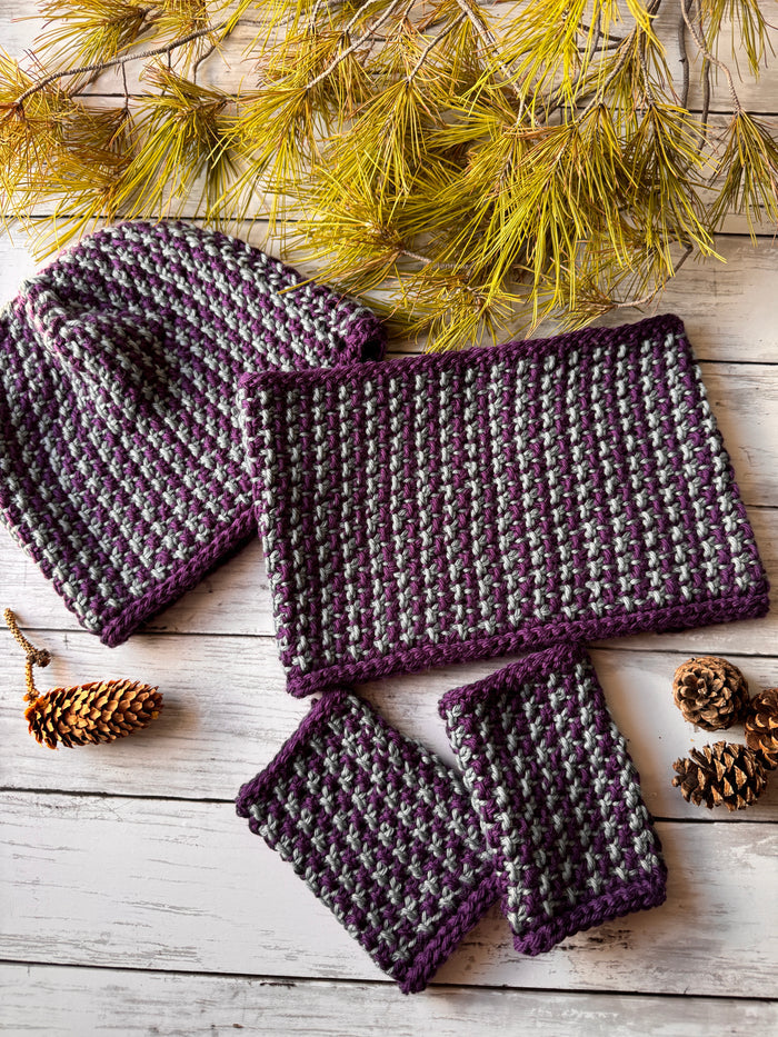 Complete Snowfall collection flat lay showing matching hat cowl and fingerless mitts in purple and gray Tunisian crochet colorwork on whitewashed wood with pine and pinecones