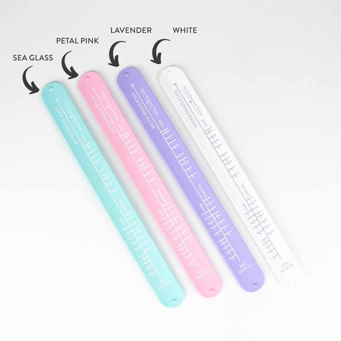 Four Sock Sizing Ruler bracelet rulers fanned out showing all available colors: Sea Glass, Petal Pink, Lavender, and White with labeled arrows pointing to each color