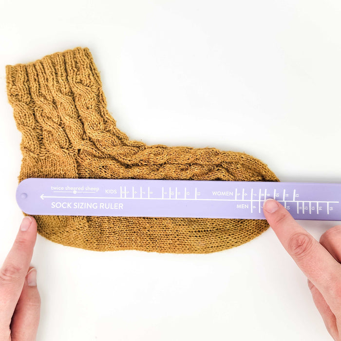Hands holding a lavender Sock Sizing Ruler against a golden cable-knit sock in progress to measure the foot length with Kids, Women, and Men size markings visible