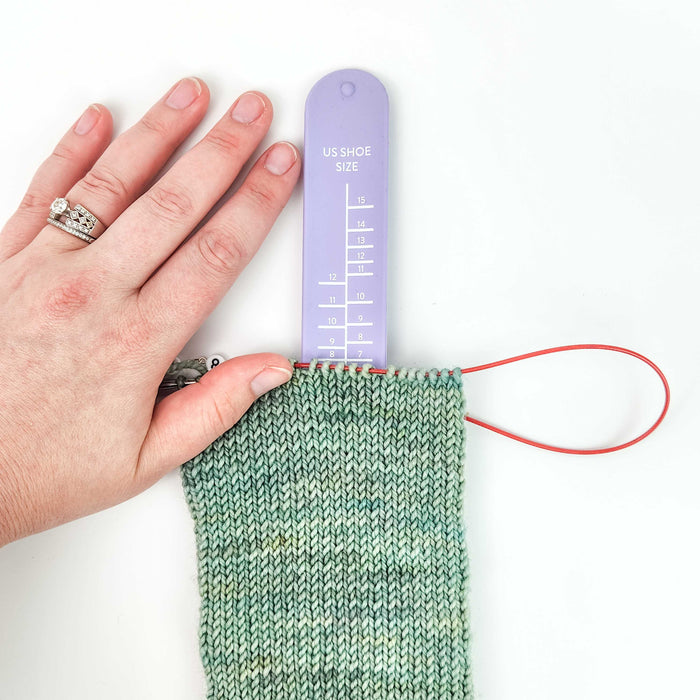 Hand holding a lavender Sock Sizing Ruler inside a mint green stockinette sock in progress on circular needles demonstrating how to measure for the correct shoe size