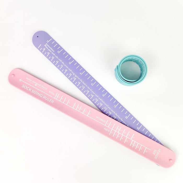 Three Sock Sizing Rulers displayed flat and curled: a pink ruler flat, a lavender ruler angled, and a sea glass ruler coiled into its bracelet form showing the slap-bracelet design