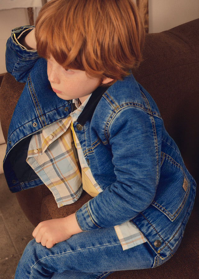 Child wearing a denim jacket and plaid shirt sitting on a brown couch.