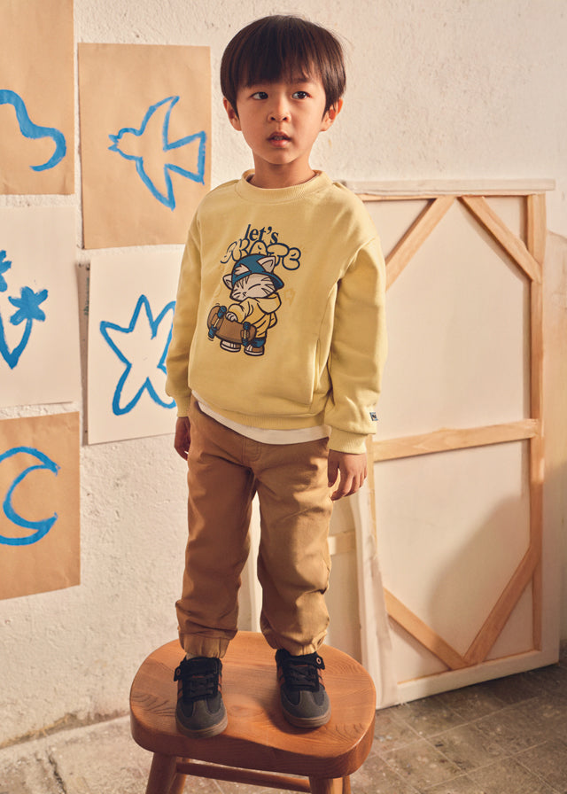 Child wearing a yellow sweater with a cartoon design, standing on a wooden stool against a wall with colorful shapes.
