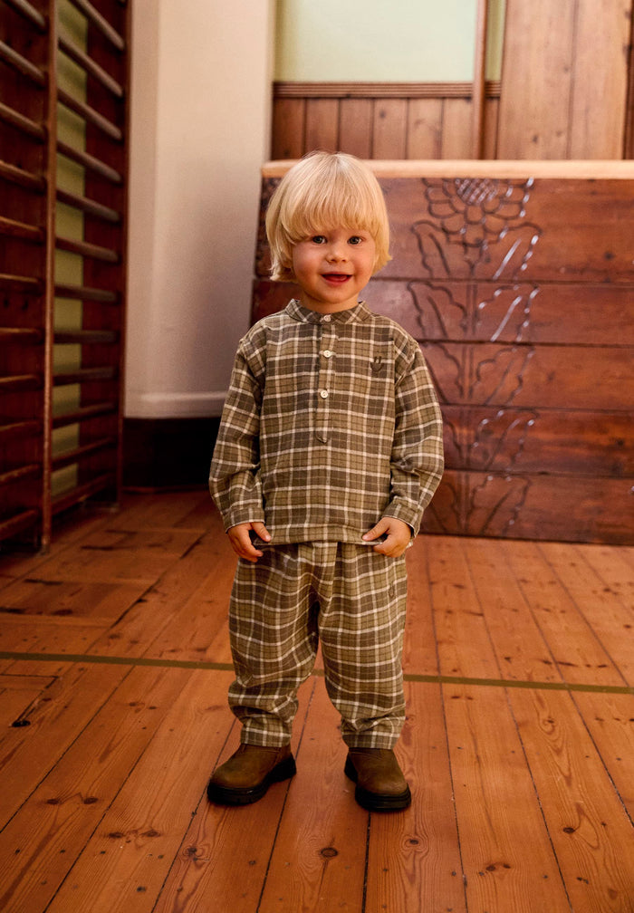Child wearing a plaid outfit standing on a wooden floor with a wooden cabinet in the background.