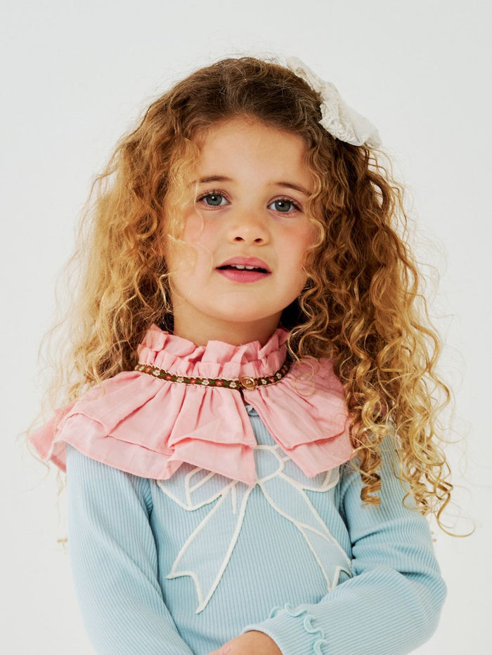 Young girl with curly hair wearing a light blue sweater and pink ruffled collar against a white background