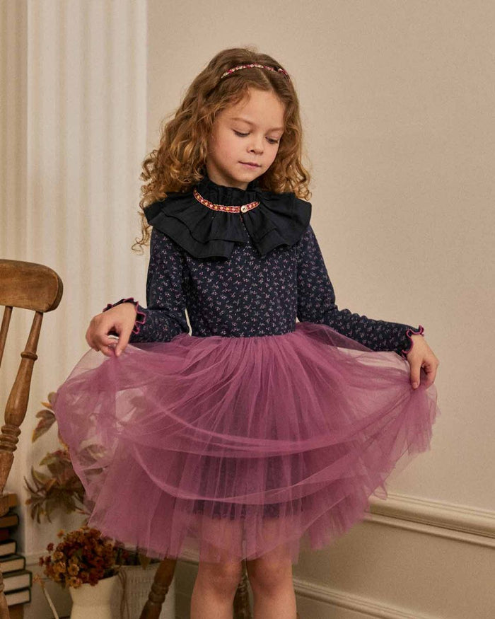 Young girl wearing a navy top with ruffled collar and a pink tulle skirt, standing indoors.