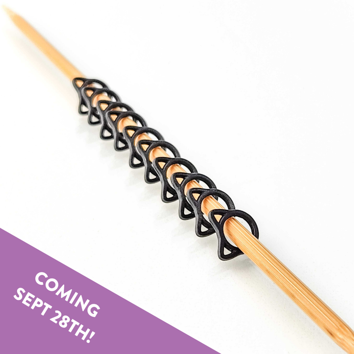 Row of black cat-shaped snag free stitch markers lined up on a bamboo knitting needle, with a Coming Sept 28th banner
