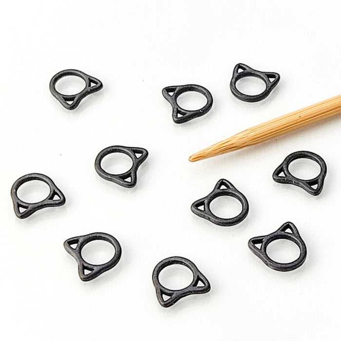 Nine black cat shaped stitch markers scattered on a white background next to a bamboo knitting needle showing size scale