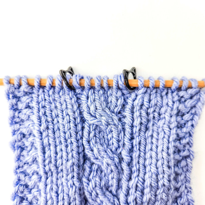 Two black cat stitch markers in use on a blue cable knit swatch on bamboo needles marking pattern sections