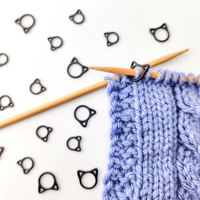 Multiple black cat stitch markers scattered on white background with bamboo needles and blue cable knit swatch showing markers in use