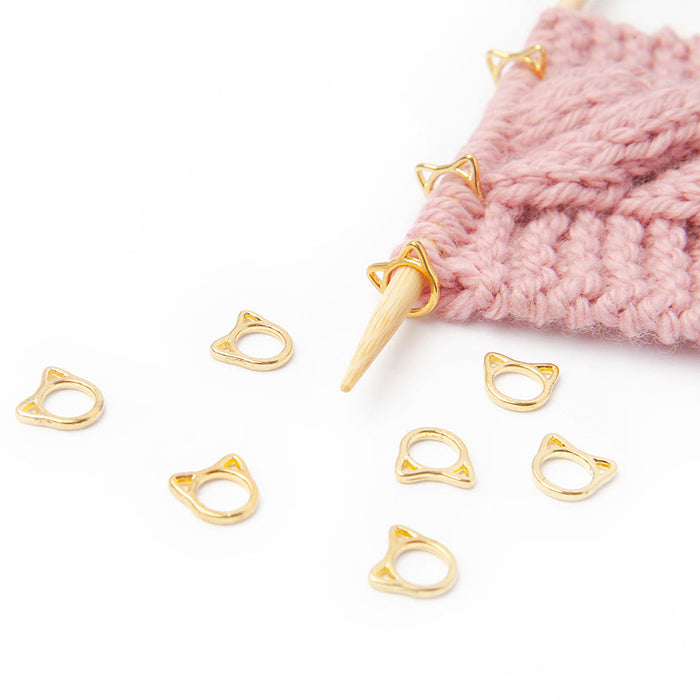 Gold cat-face ring stitch markers scattered beside pink knitting on bamboo needle with markers in use