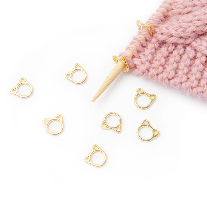 Eight gold cat-face ring stitch markers scattered on white background beside pink knitting on bamboo needle