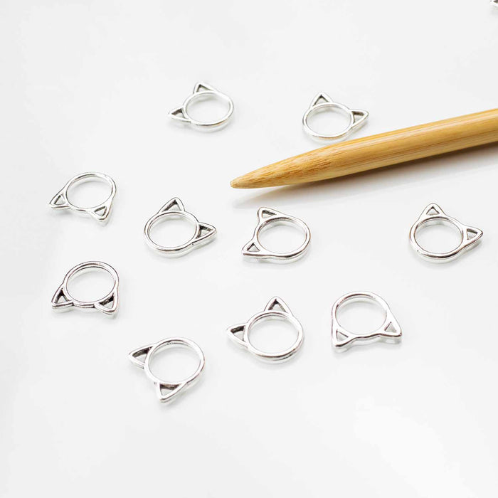 Eight silver cat-face ring stitch markers scattered on white background with bamboo knitting needle