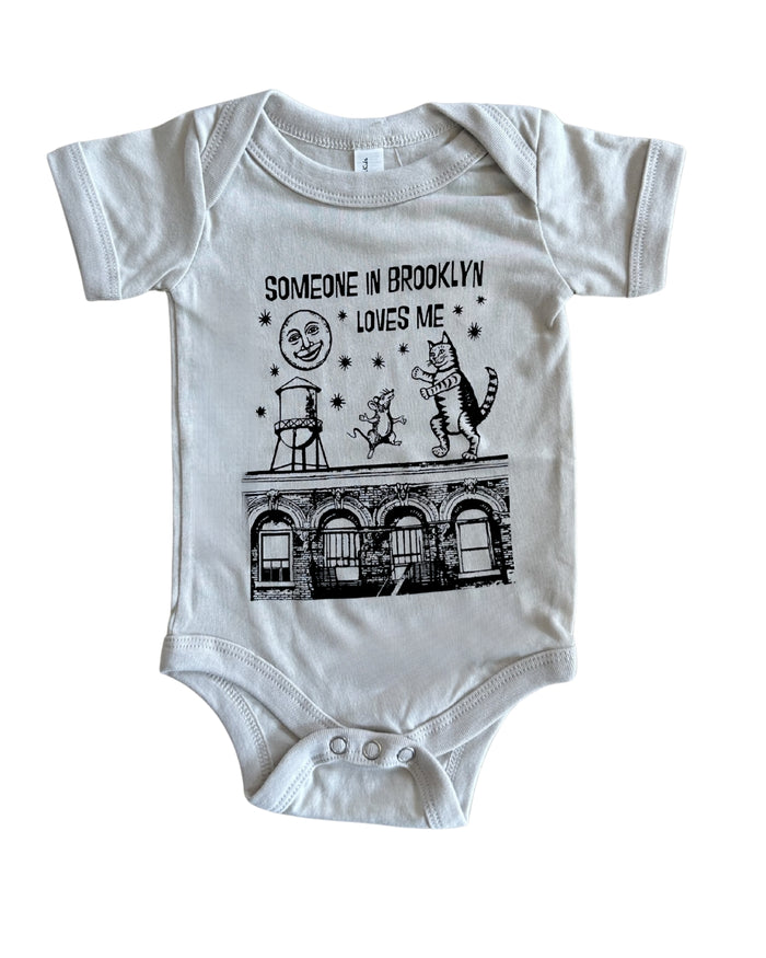 A grey short-sleeve baby bodysuit with a black graphic print on the front. The graphic features the text 'SOMEONE IN BROOKLYN LOVES ME' above a whimsical illustration of a smiling moon, a figure on a ladder, a cat, stars, and a building facade.