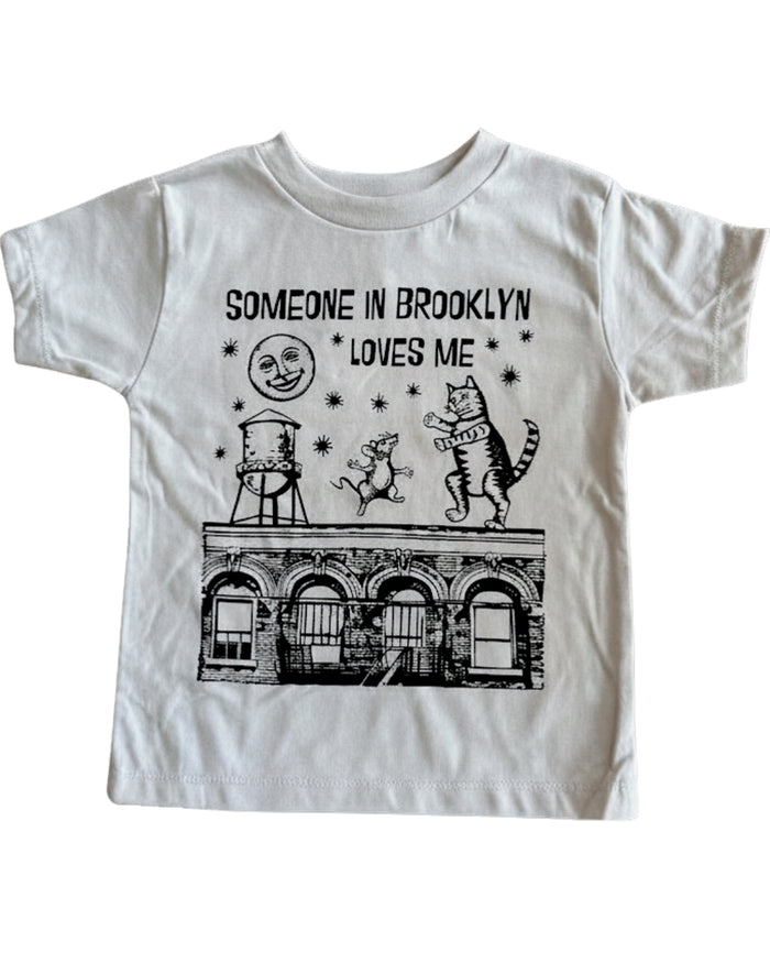 A grey short-sleeve crewneck t-shirt featuring a black graphic print of a Brooklyn building, a water tower, a cat, and a rat with the text 'SOMEONE IN BROOKLYN LOVES ME'.