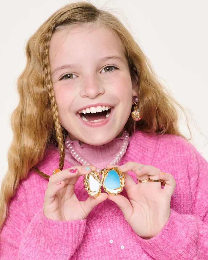 Young girl holding a decorative mirror with a blue and gold frame, wearing a pink sweater and pearl necklace.