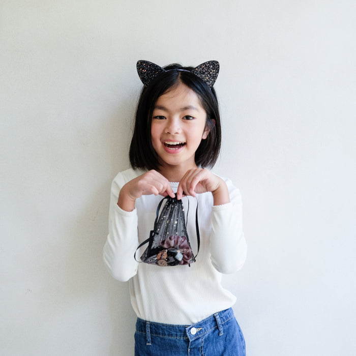 A young girl with black cat ears headband smiling and holding a small black glitter drawstring bag with pink and white cat illustrations, wearing a white long-sleeve shirt and blue jeans against a plain white wall.