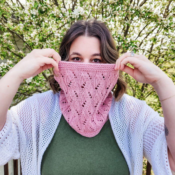 Woman holding up pink lace knit Spring Vibes Cowl showing leaf lace pattern detail with flowering tree behind