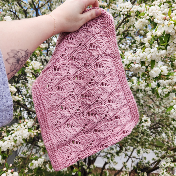 Hand holding pink lace knit Spring Vibes Cowl showing full leaf lace stitch pattern against flowering tree backdrop