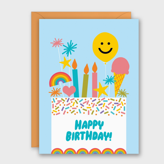 A colorful birthday card with a light blue border and an orange envelope behind it. The card features a white lower section with the words “HAPPY BIRTHDAY!” in blue, decorated with illustrated candles, balloons, fireworks, a rainbow, stars, and sprinkles.