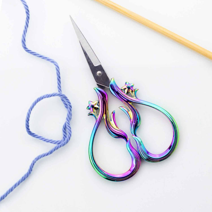 Star Dust Rainbow embroidery scissors closed on a white surface with blue yarn and a bamboo knitting needle showing the iridescent rainbow flame handles