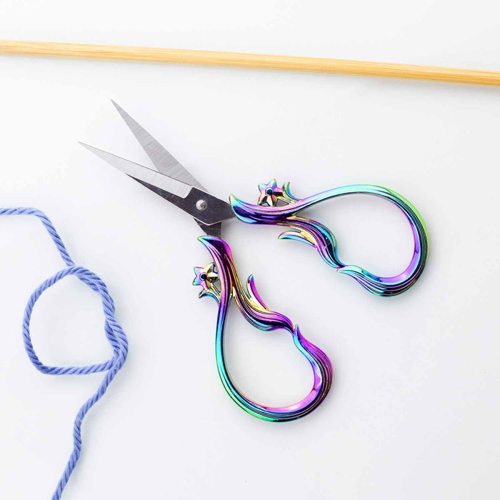 Star Dust Rainbow embroidery scissors partially open on a white surface with blue yarn and bamboo knitting needle showing the sharp pointed stainless steel blades