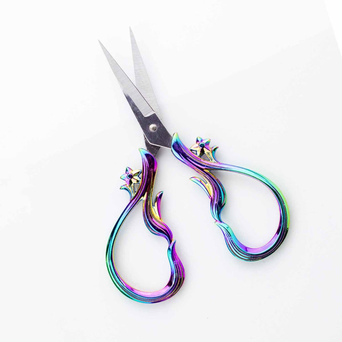 Star Dust Rainbow embroidery scissors shown open at an angle on a white surface with blue yarn and bamboo knitting needle