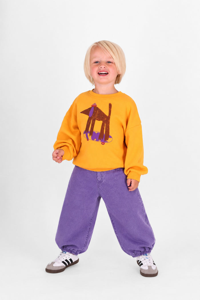 A young child with blonde hair stands against a white studio background wearing a bright yellow sweatshirt with a brown and purple dog graphic, loose purple pants, and white sneakers.