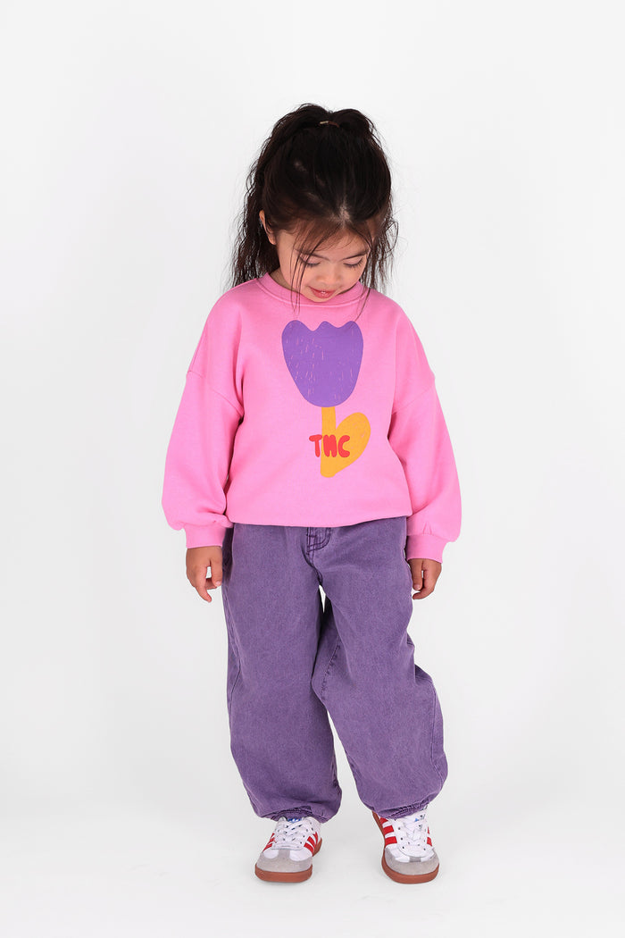 A young girl wearing a pink sweatshirt with a purple tulip graphic and the letters 'THC' in yellow, paired with loose purple pants and white sneakers, standing against a plain white background.