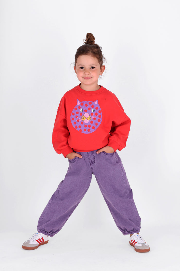 A young girl with her hair in a bun stands in a studio setting, wearing a bright red sweatshirt with a purple polka-dot cat face graphic on the front, paired with loose purple corduroy pants and white sneakers with red stripes.