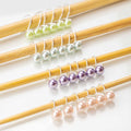 Four sets of infinity ring style pastel pearl stitch markers in green, silver, purple, and pink on bamboo knitting needles