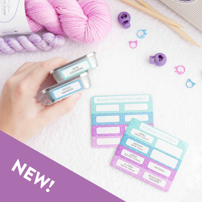 Hand holding two stacked storage tins labeled Row Counters and Stitch Markers with pre-printed and blank label sticker sheets in pastel pink, blue, and purple alongside yarn and knitting accessories
