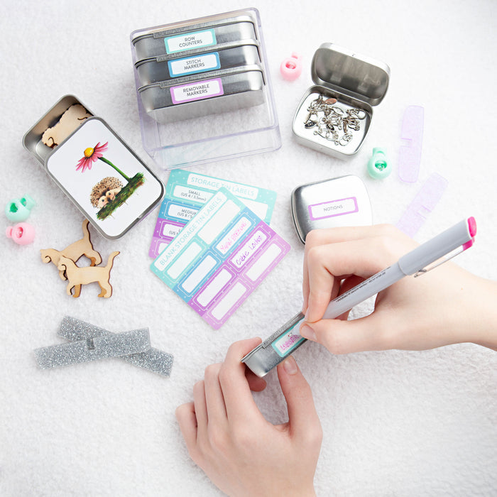 Hands writing on a blank storage tin label sticker with a marker pen surrounded by labeled tins, sticker sheets, hedgehog tin, wooden cat figures, and knitting notions