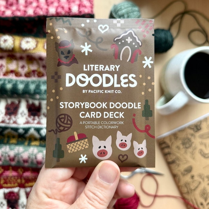 Hand holding the brown Literary Doodles Storybook Doodle Card Deck pouch, printed with pig, gingerbread house and wolf motifs, above a striped colorwork knit swatch, yarn balls and a cup of coffee