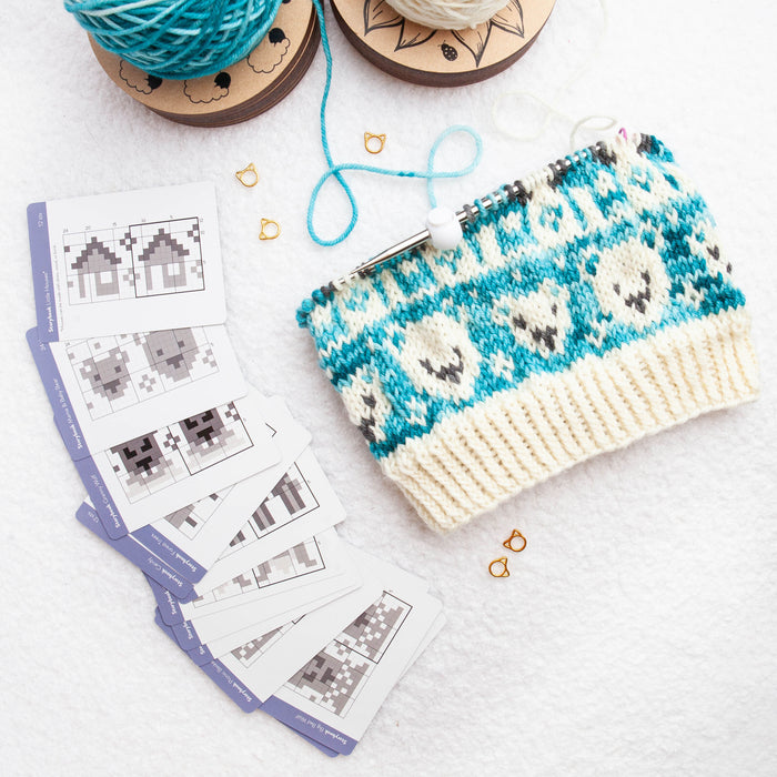 Teal and cream colorwork knitting in progress on a circular needle showing bear motifs, beside a fanned stack of Storybook Doodle Deck chart cards, two yarn cakes and gold stitch markers