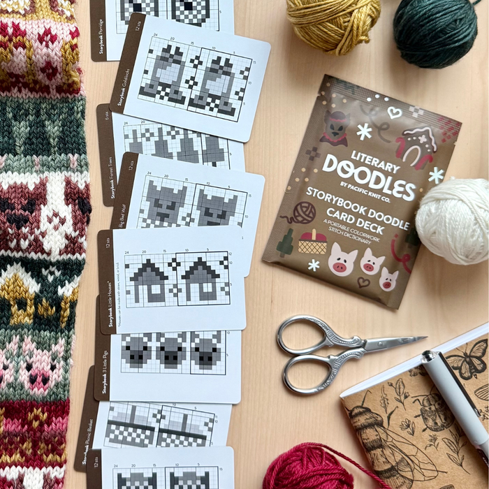 Storybook Doodle Deck chart cards fanned across a wood table next to the brown deck pouch, yarn balls, embroidery scissors and a knit colorwork swatch