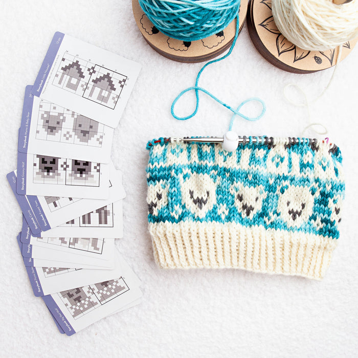 Cream and teal colorwork knit cowl in progress with bear motifs on a circular needle, next to a fan of Storybook Doodle Deck pattern cards and two yarn cakes on a white background