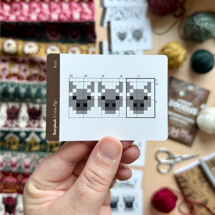 Hand holding the Storybook 3 Little Pigs chart card from the Storybook Doodle Deck, showing three pig colorwork charts, with yarn and a knit swatch blurred behind