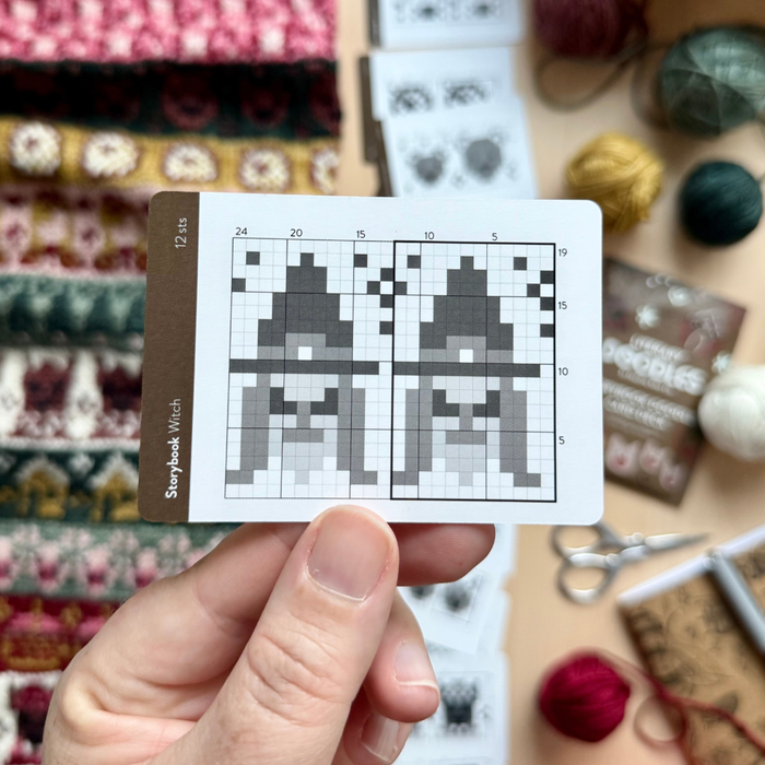 Hand holding the Storybook Witch chart card from the Storybook Doodle Deck, showing two witch colorwork charts, with yarn balls and a colorwork swatch blurred behind