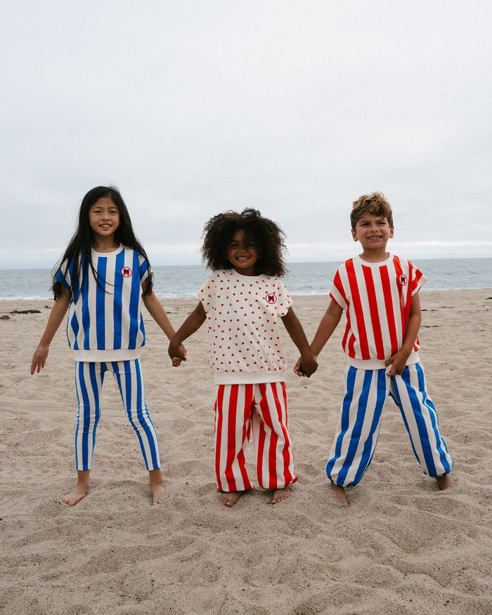 Three children holding hands on a beach wearing matching outfits with blue, white, and red stripes.
