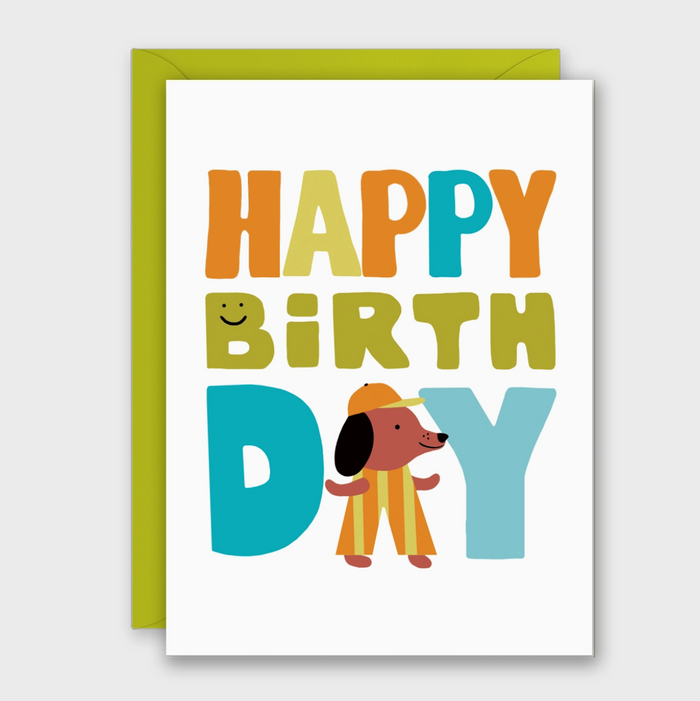 A white birthday card with a bright green envelope behind it. The card reads “HAPPY BIRTH DAY” in large multicolored letters, with a cartoon dog wearing a yellow hat and striped pants standing between within the word “DAY".
