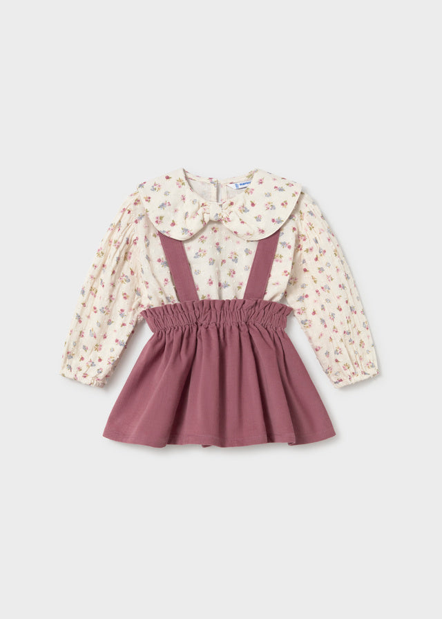 Children's outfit with floral blouse and purple pinafore on a light gray background